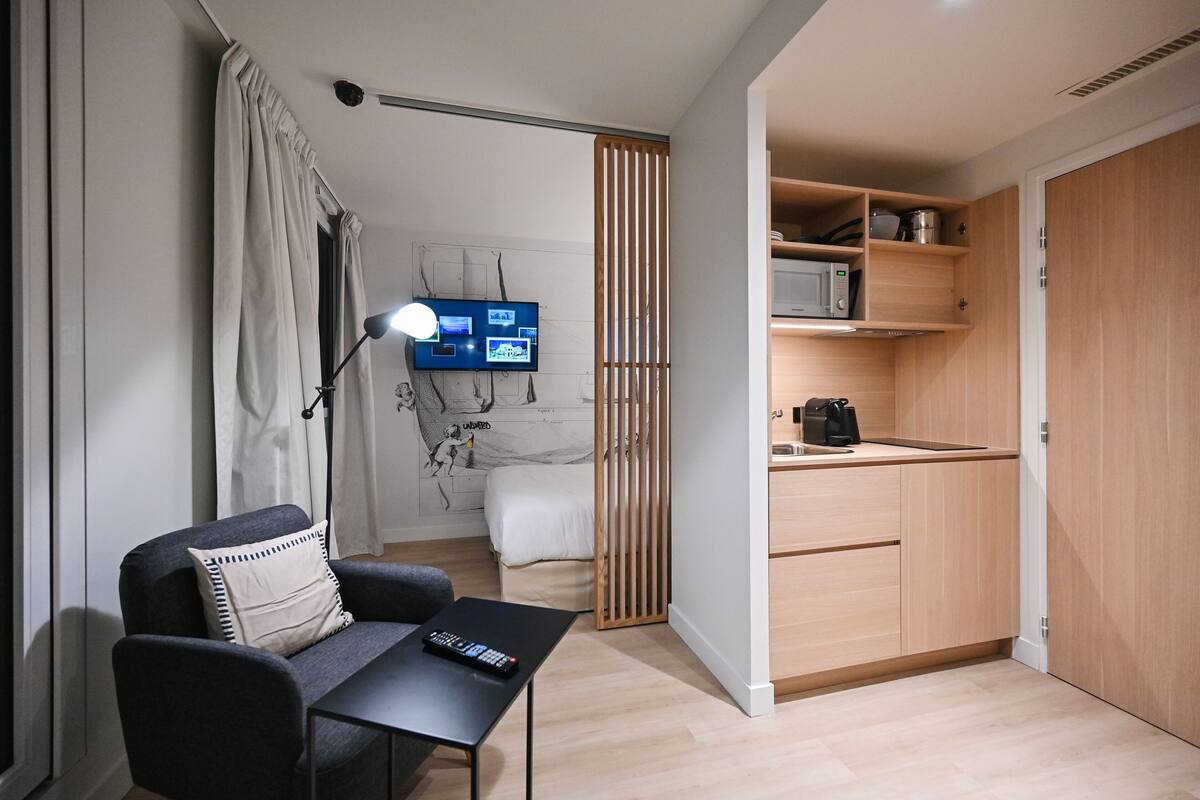 A compact room with a bed, a desk with a lamp, a TV, and a kitchenette.