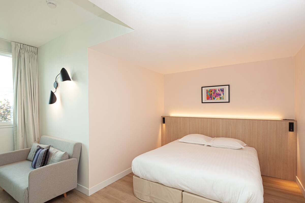 A modern hotel room with a bed, a sofa, a wall-mounted lamp, and a framed picture.
