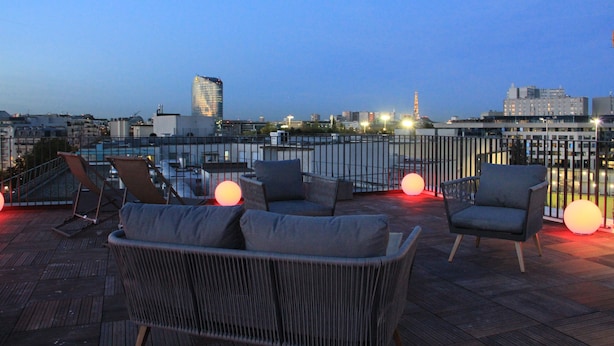 A rooftop terrace with wicker furniture and red spherical lights, overlooking a cityscape with buildings and a tower. - Finestate Coliving Mairie d'Issy  (Issy-les-Moulineaux)