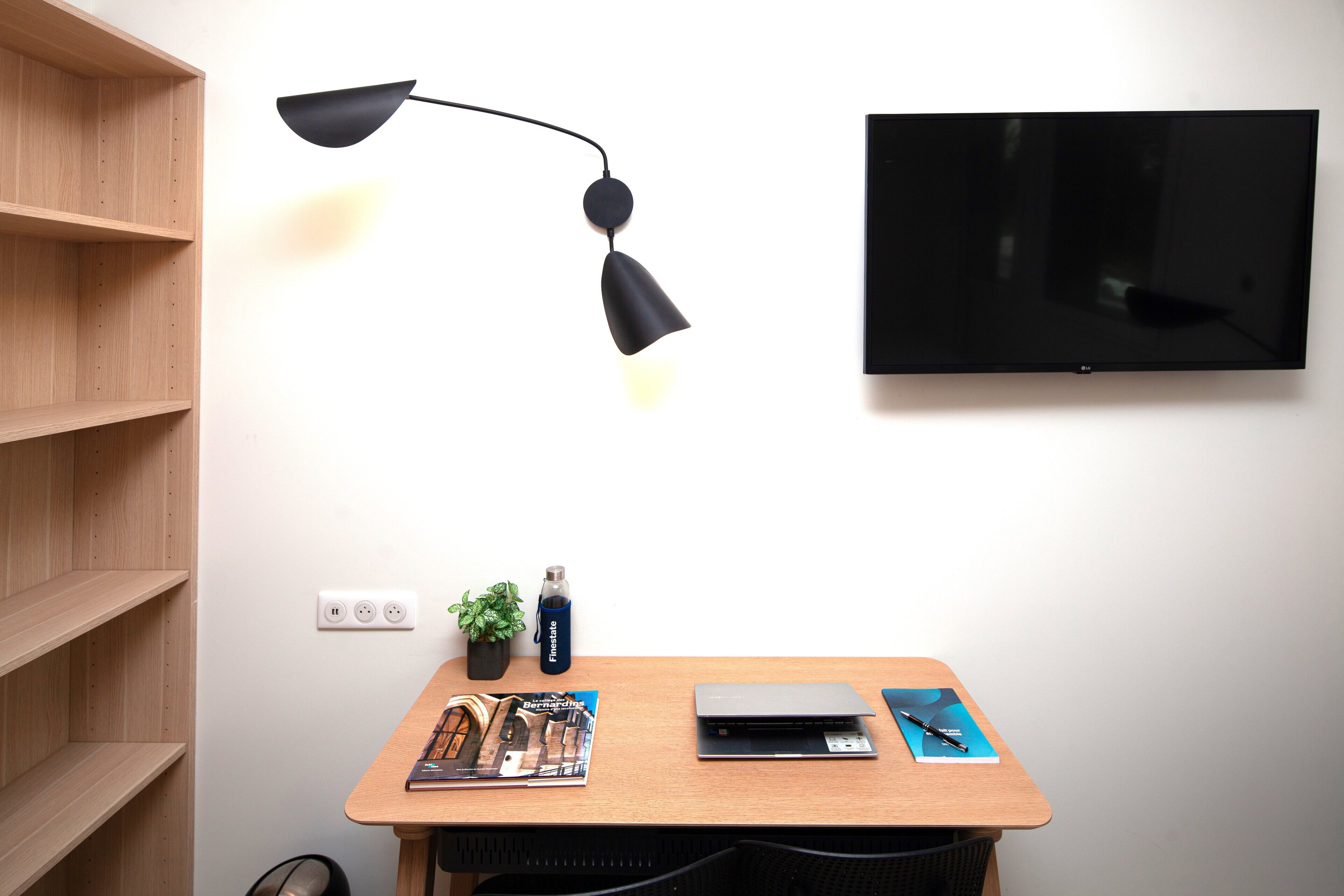 Ett rum med ett skrivbord i trä, en bärbar dator, en tidning, en växt, en vattenflaska, en lampa och en tv som är monterad på väggen.