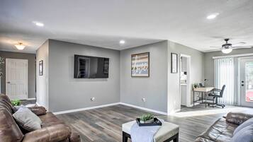 A spacious living room with a large flat-screen TV, a dining area with a desk and chair, and a ceiling fan.