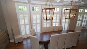A dining room with a large wooden table surrounded by white upholstered chairs, two hanging lantern-style lights, and white shutters on the windows. - Beach luxury at it's best!! (Rehoboth Beach)