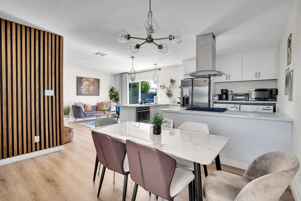 A modern kitchen with white cabinets, a stainless steel refrigerator, and a built-in oven. There is a dining area with a white table and two chairs. - Minnie's Marvelous Escape 8 minutes to Disney & Convention Center (Anaheim)