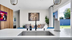 Une cuisine moderne dotée d’un grand îlot central, d’un téléviseur fixé au mur diffusant des services de streaming, d’un tableau représentant un lion et donnant sur un jardin.