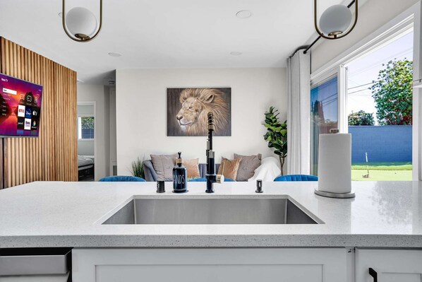 A modern kitchen with a large island, a mounted television displaying streaming services, a painting of a lion, and a view of a backyard. - Minnie's Marvelous Escape 8 minutes to Disney & Convention Center (Anaheim)