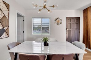 Un comedor con una mesa de mármol blanco, sillas de cuero marrón, una araña, una ventana con persianas y una puerta de madera.