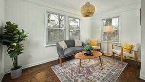 En stue med en grå sofa, et rundt sofabord og en gulvlampe. Der er en stor plante i en grå potte og et mønstret tæppe på gulvet.