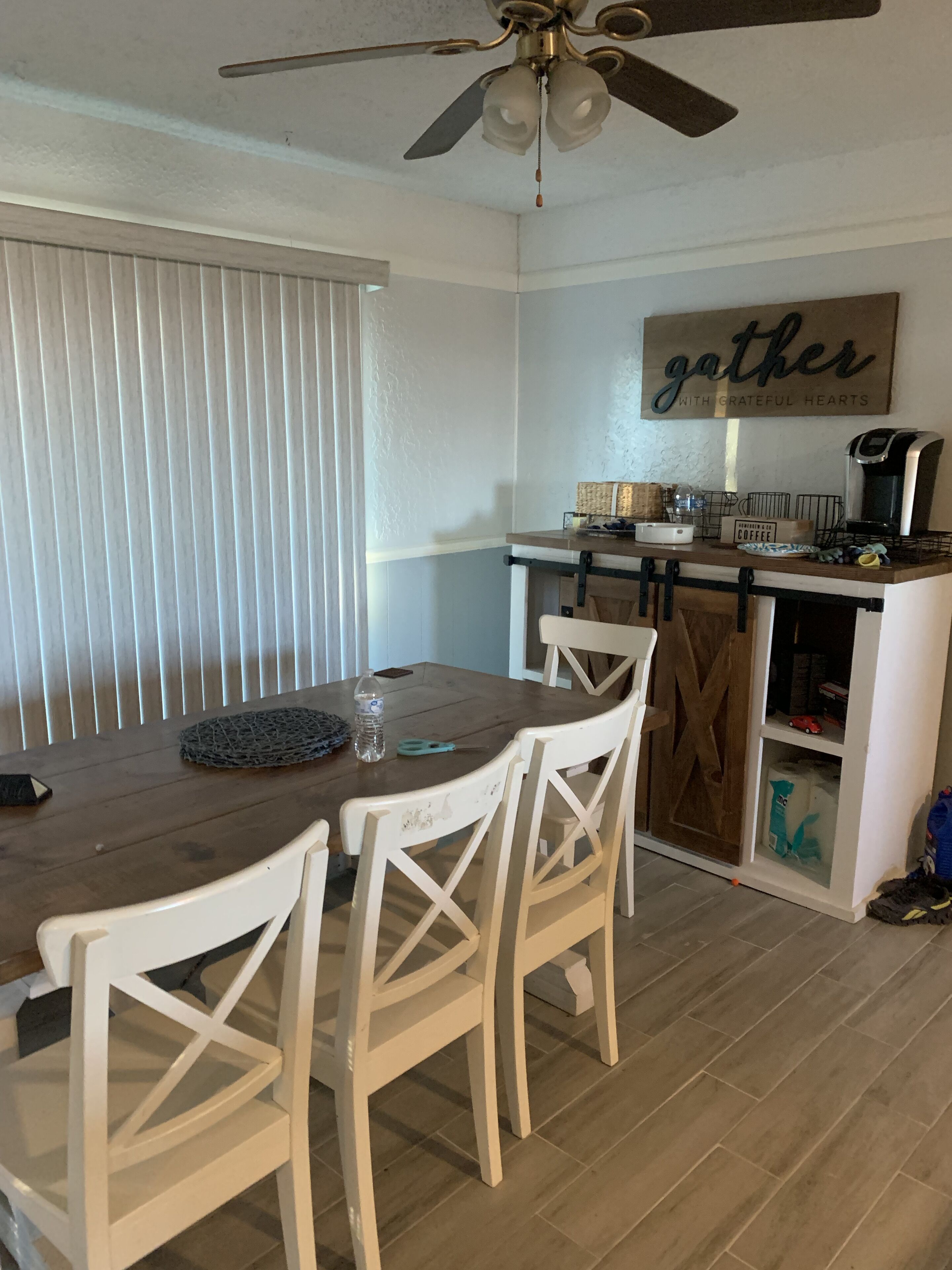 A dining area with a wooden table and white chairs, a ceiling fan, a kitchen area with a coffee maker, and a sign that says "gather with grateful hearts".