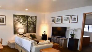 A modern living room with a sofa, a TV, a coffee table, and a dining table. There are framed pictures on the wall and a large window. - 5 Star Upscale Unit Near Canyon Lodge (Mammoth Lakes)