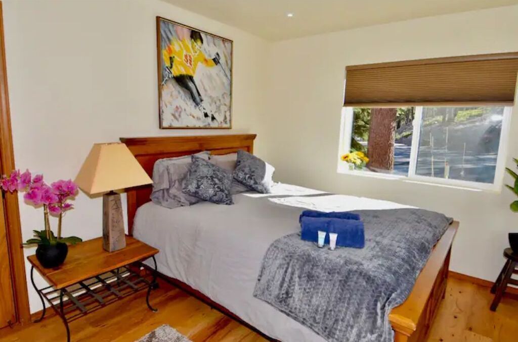 A bedroom with a bed, a nightstand, a lamp, a painting, a window with a view, and a plant.