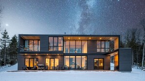 Une maison moderne à deux étages, dotée de grandes fenêtres et d’un balcon, entourée de neige.