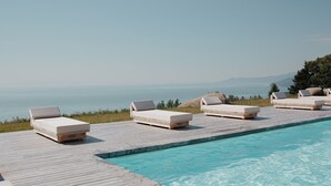 Un espace aménagé au bord de la piscine, avec des chaises longues et une terrasse en bois donnant sur une étendue d’eau.