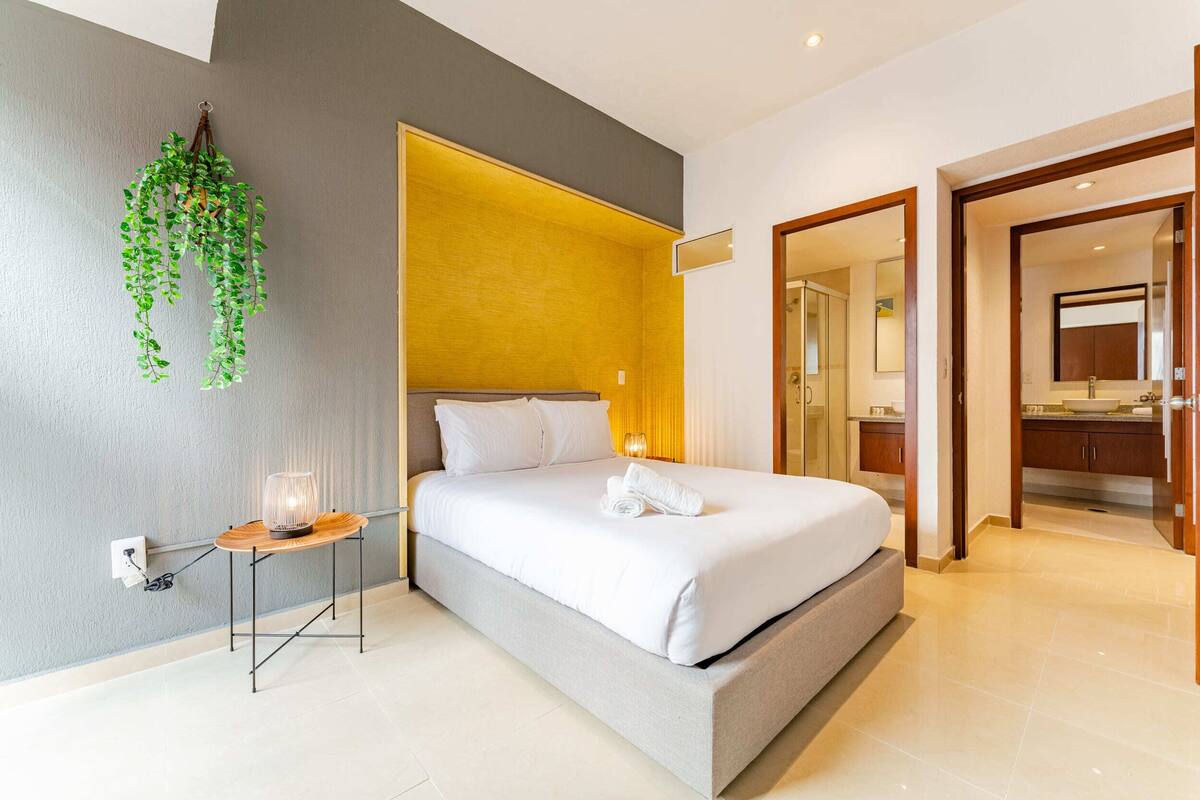 Ein modernes Hotelzimmer mit einem großen Bett, einem Nachttisch mit Lampe, einer hängenden Pflanze und einem durchgegangenen Tür erkennbaren Badezimmer.