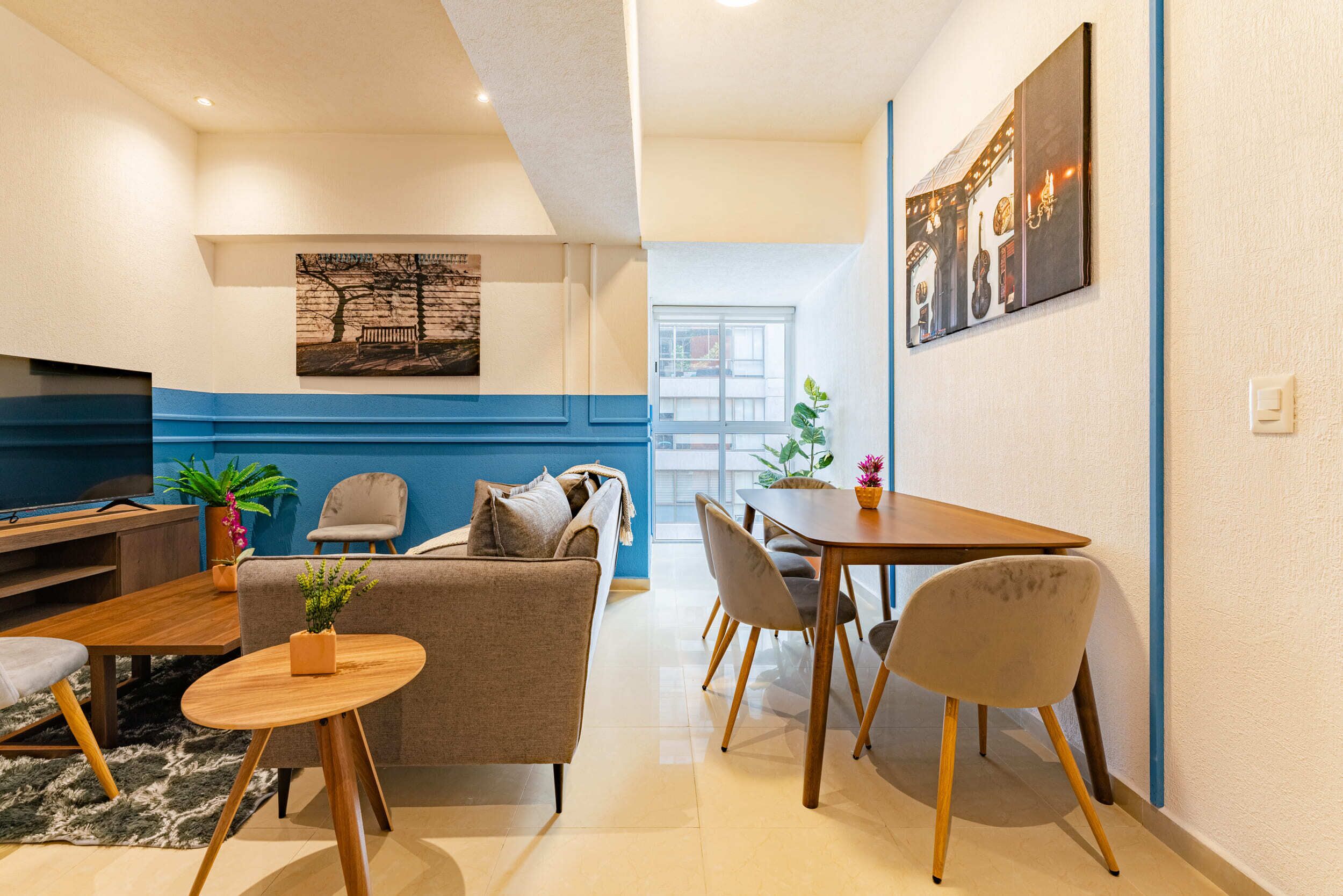 A modern living room with a sofa, chairs, a coffee table, and a dining table. There is a TV, a plant, and artwork on the walls.