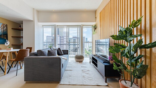 A modern living room with a grey sofa, a dining area with wooden chairs, a large window with a city view, and a potted plant. - Anahuac Apartments by VH (Mexico City)