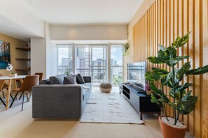 Una sala de estar moderna con un sofá gris, un comedor con sillas de madera, un ventanal con vistas a la ciudad y una planta en maceta.