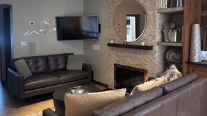 Una sala de estar moderna con un amplio sofá seccional marrón oscuro, un televisor de pantalla plana montado en la pared, una chimenea con un espejo encima y una estantería llena de libros y objetos decorativos.