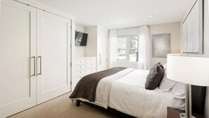 Un dormitorio con cama, mesita de noche, lámpara, televisión, armario y ventana con cortinas.