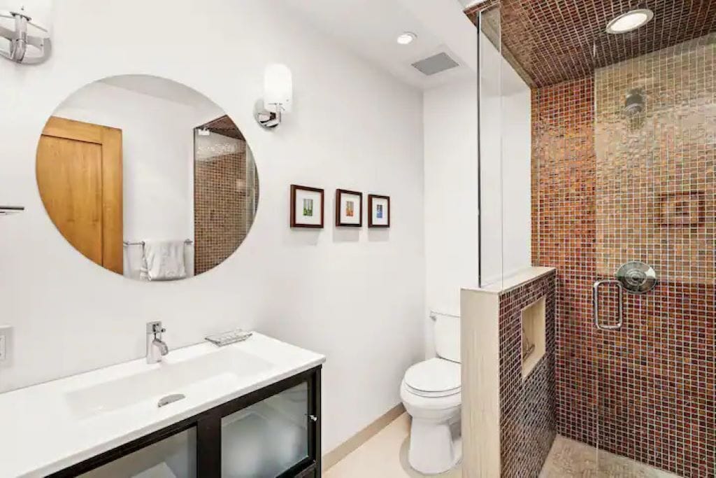 Un baño moderno con un espejo redondo, lavamanos blanco y una ducha con mosaicos.