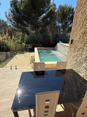 An outdoor pool area with a table and chairs, surrounded by trees and a stone wall.