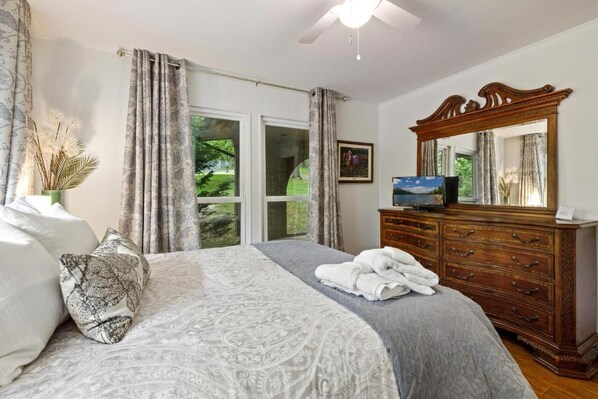 A bedroom with a large bed, a wooden dresser, a ceiling fan, and a large window with curtains.
