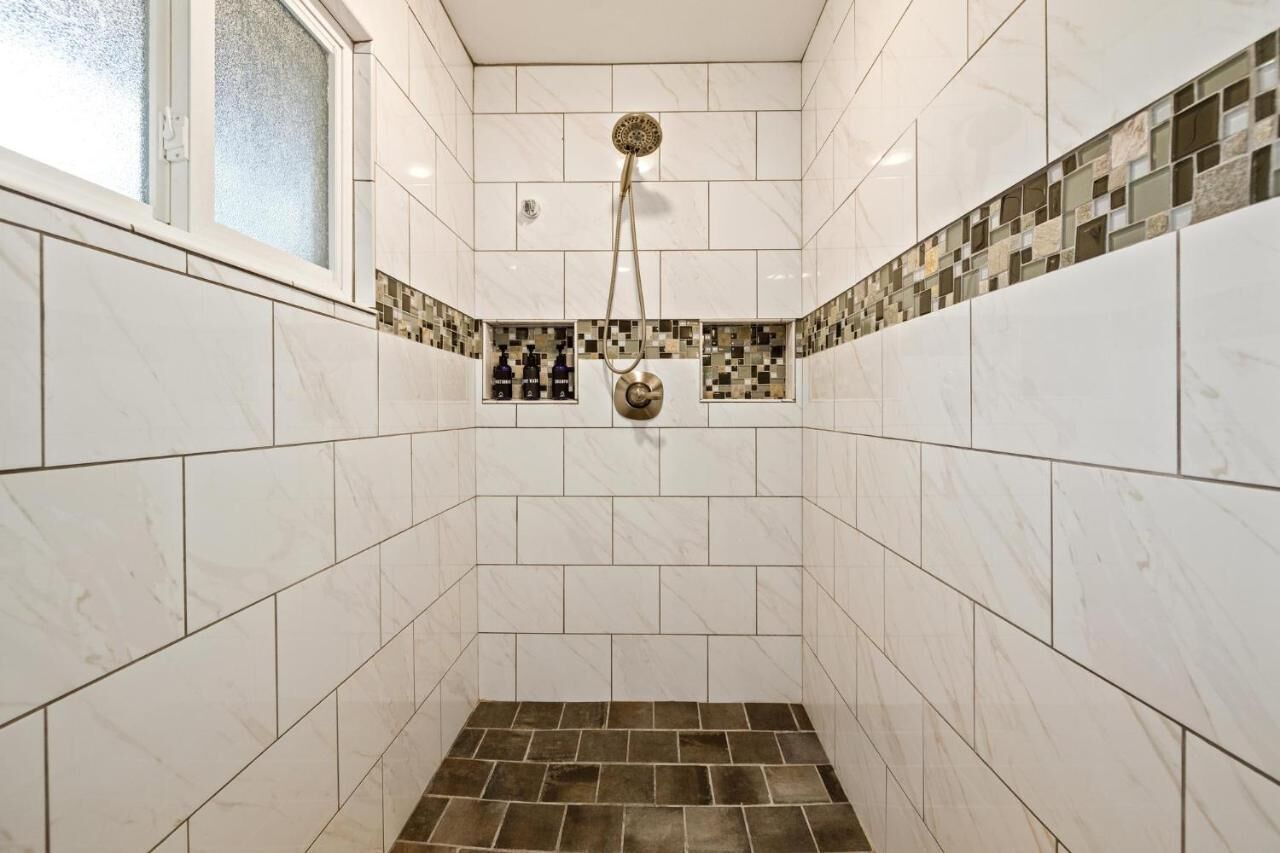 Eine Dusche mit goldfarbener Duschbrause, weißen Fliesen und einer Bordüre aus Mosaikfliesen.