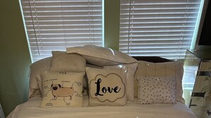 Un letto con una coperta a motivi geometrici, diversi cuscini con scritte e disegni, e una finestra con persiane sullo sfondo.