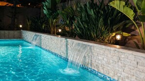 Piscina com cachoeira, cercada por plantas tropicais exuberantes e uma parede de pedra.