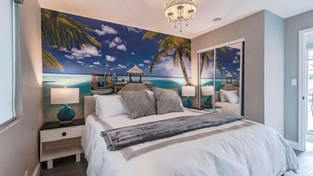 Quarto com cama grande, mesa de cabeceira, abajur e um mural na parede com cena de praia.