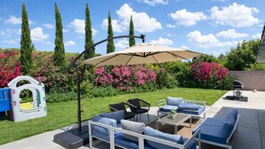 Um pátio externo com mesa e cadeiras, um grande guarda-sol e um parquinho.