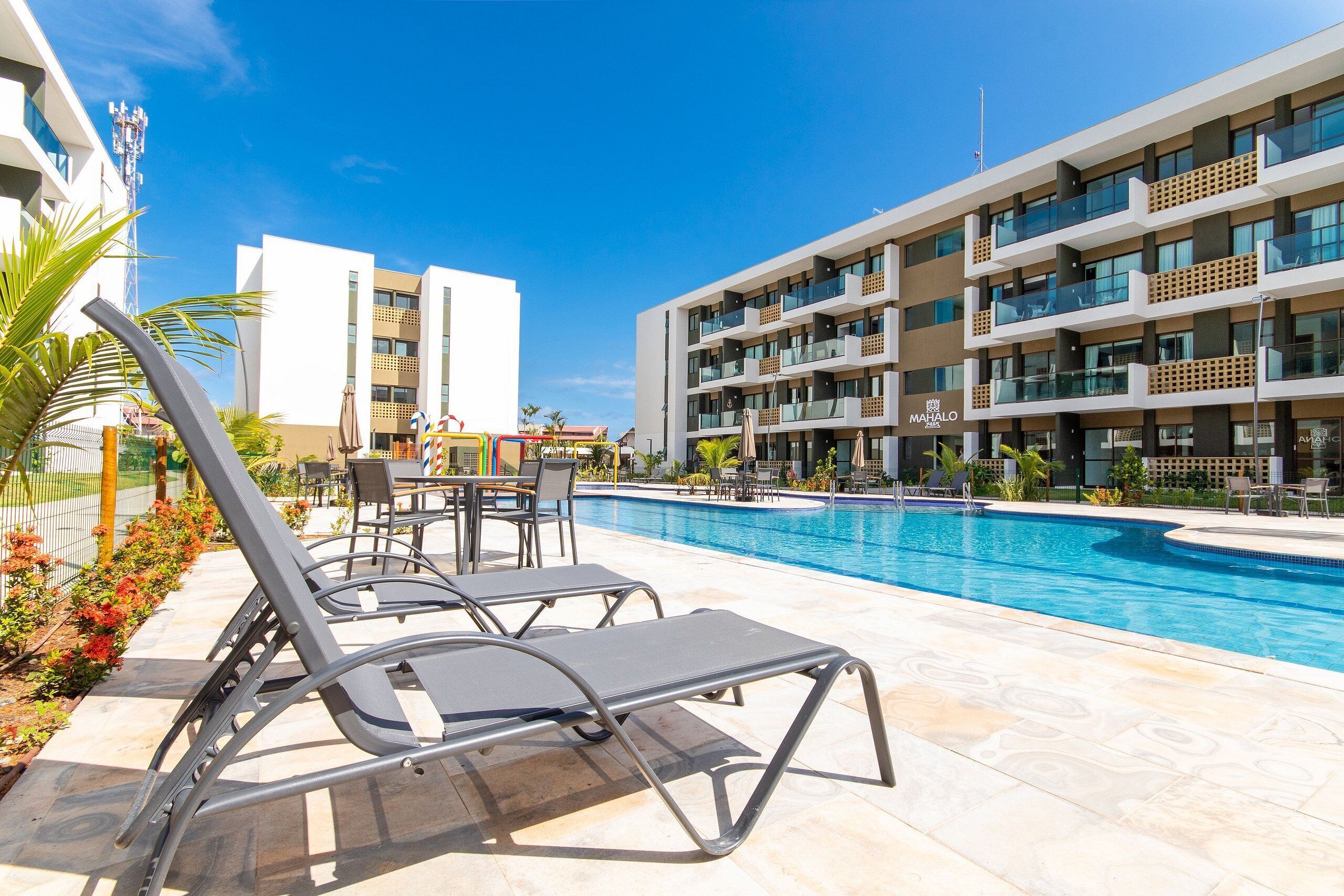 A poolside area with lounge chairs, a dining table, and a modern building with balconies.