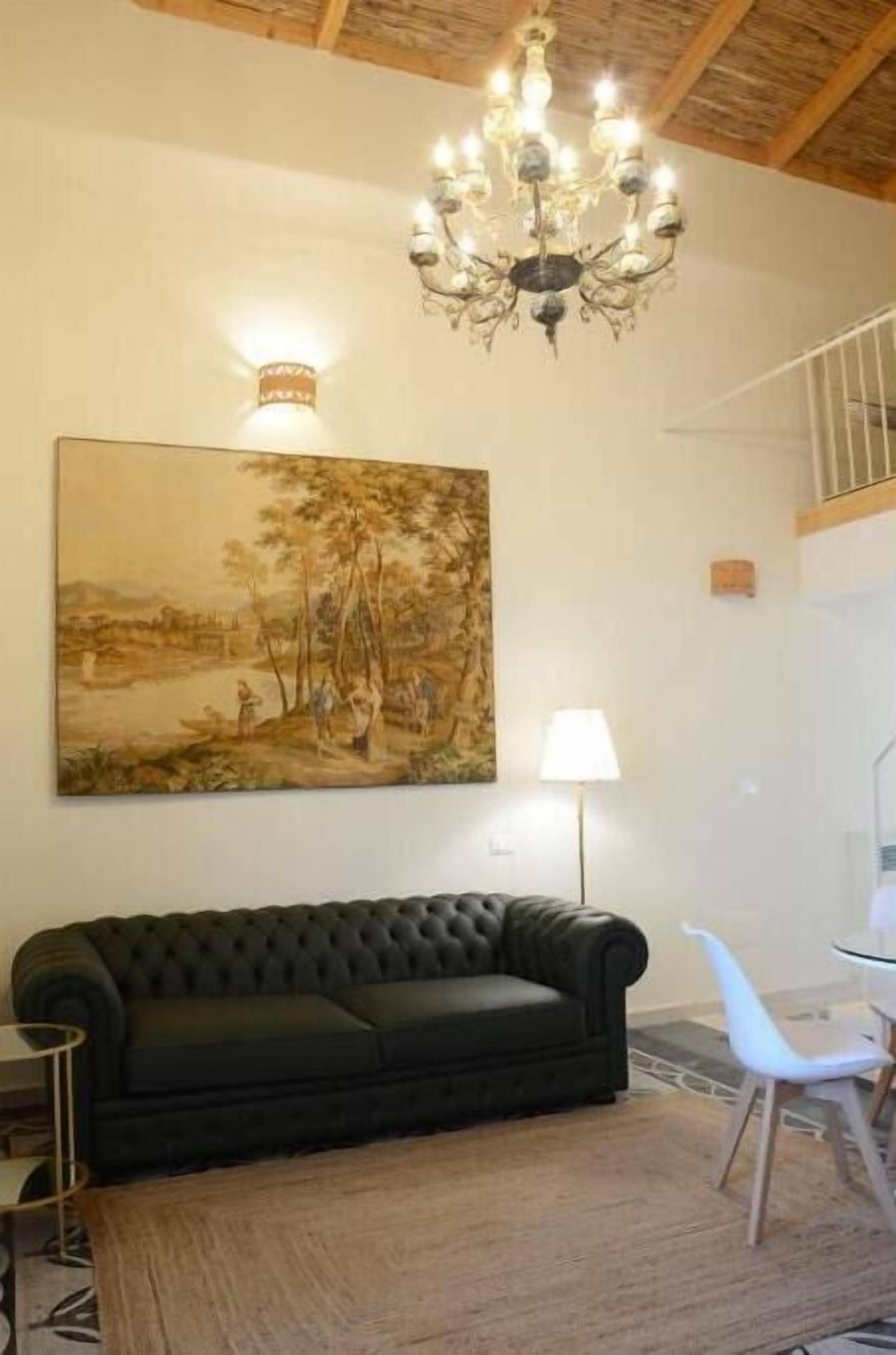 A living room with a black sofa, a white chair, a chandelier, and a large landscape painting on the wall.