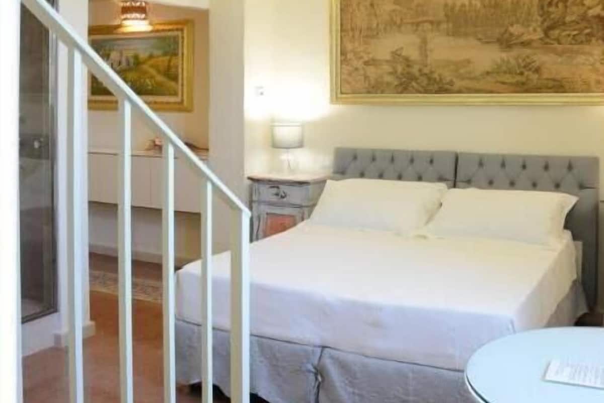 A bedroom with a large bed, a small round table, a painting on the wall, and a staircase leading to an upper level.