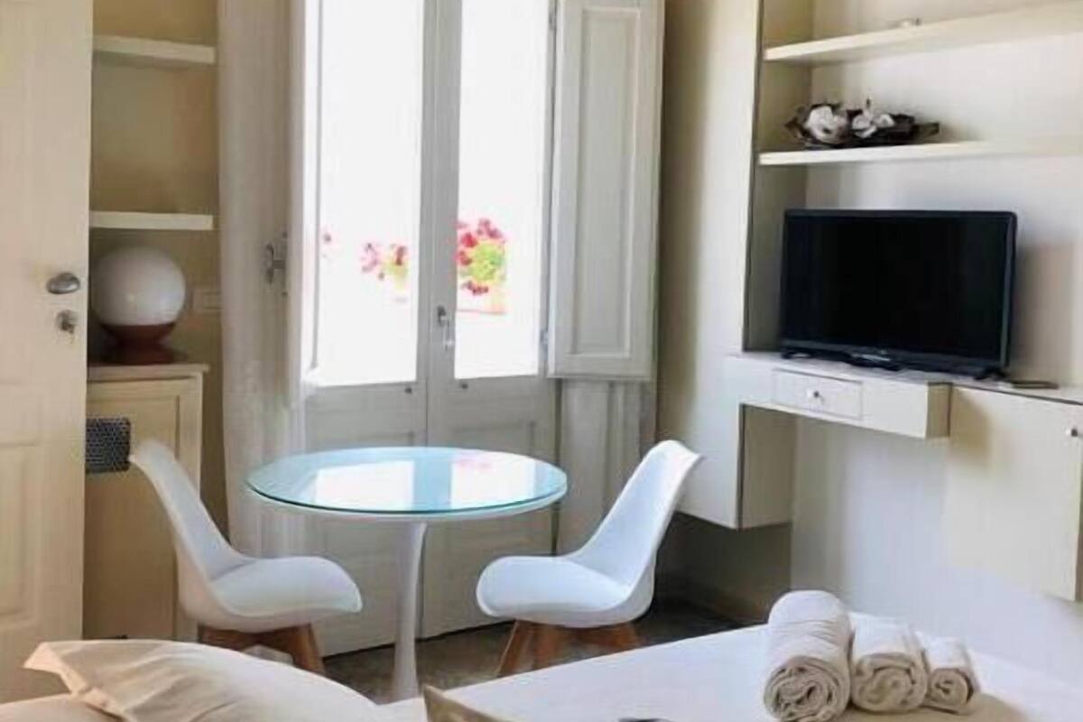 A hotel room with a bed, two white chairs, a small round table, a TV on a shelf, and a window with curtains.