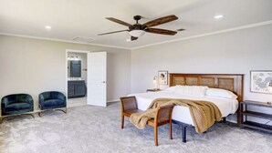 A bedroom with a bed, two chairs, a ceiling fan, a nightstand, and a door leading to a bathroom.