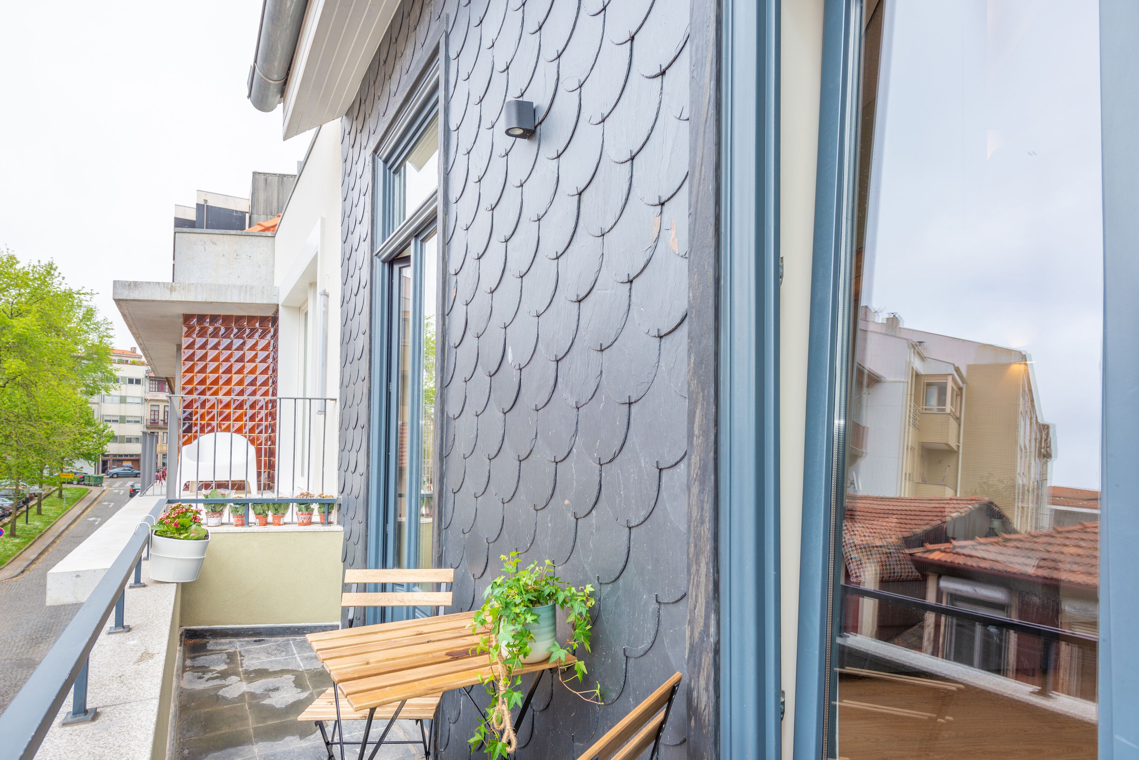 Un balcon avec un banc en bois, des plantes en pot et la façade d’un immeuble moderne ornée d’un motif original en forme d’écailles de poisson.