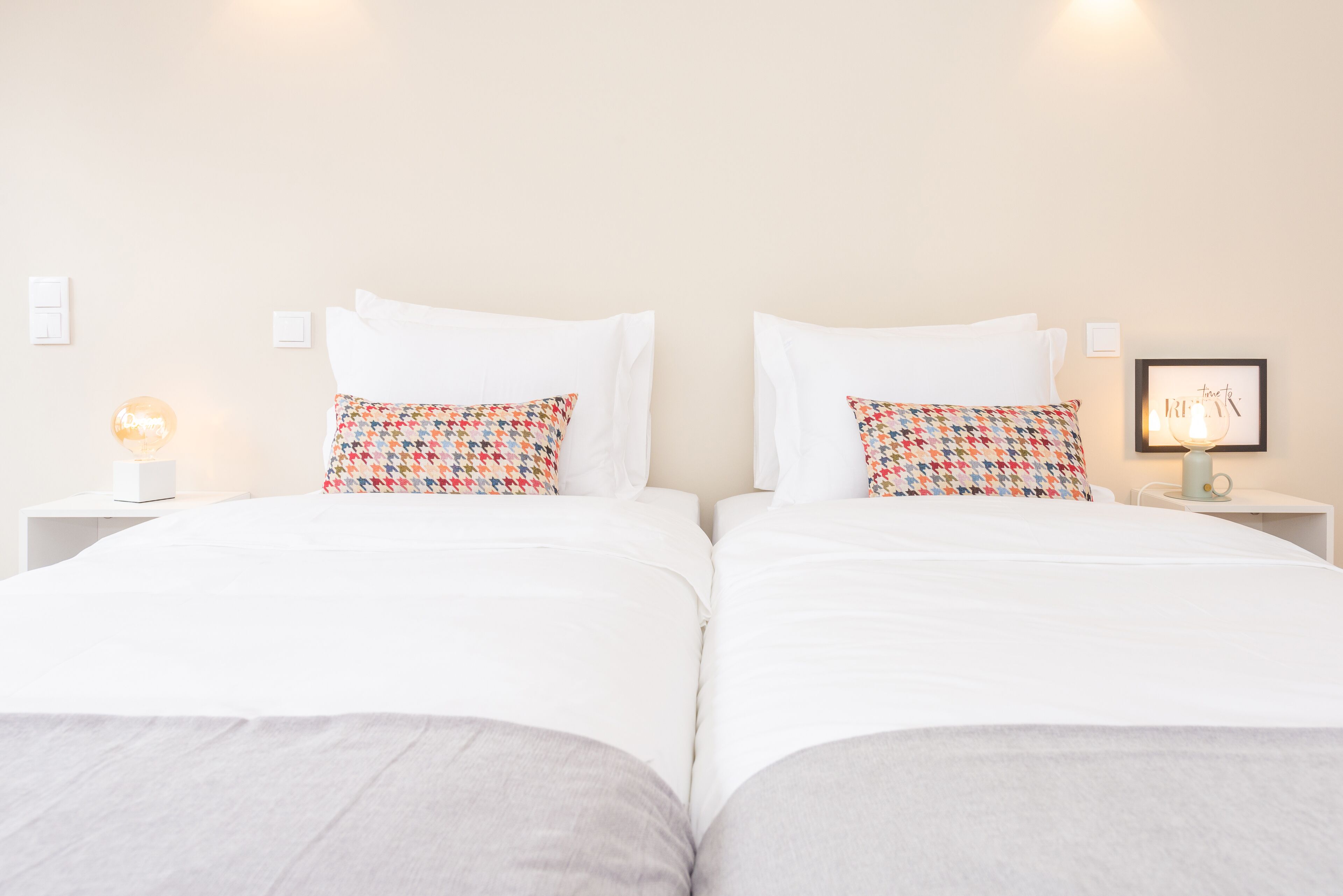 A double bed with white bedding and patterned pillows, a bedside table with a lamp, and a framed picture on the wall.
