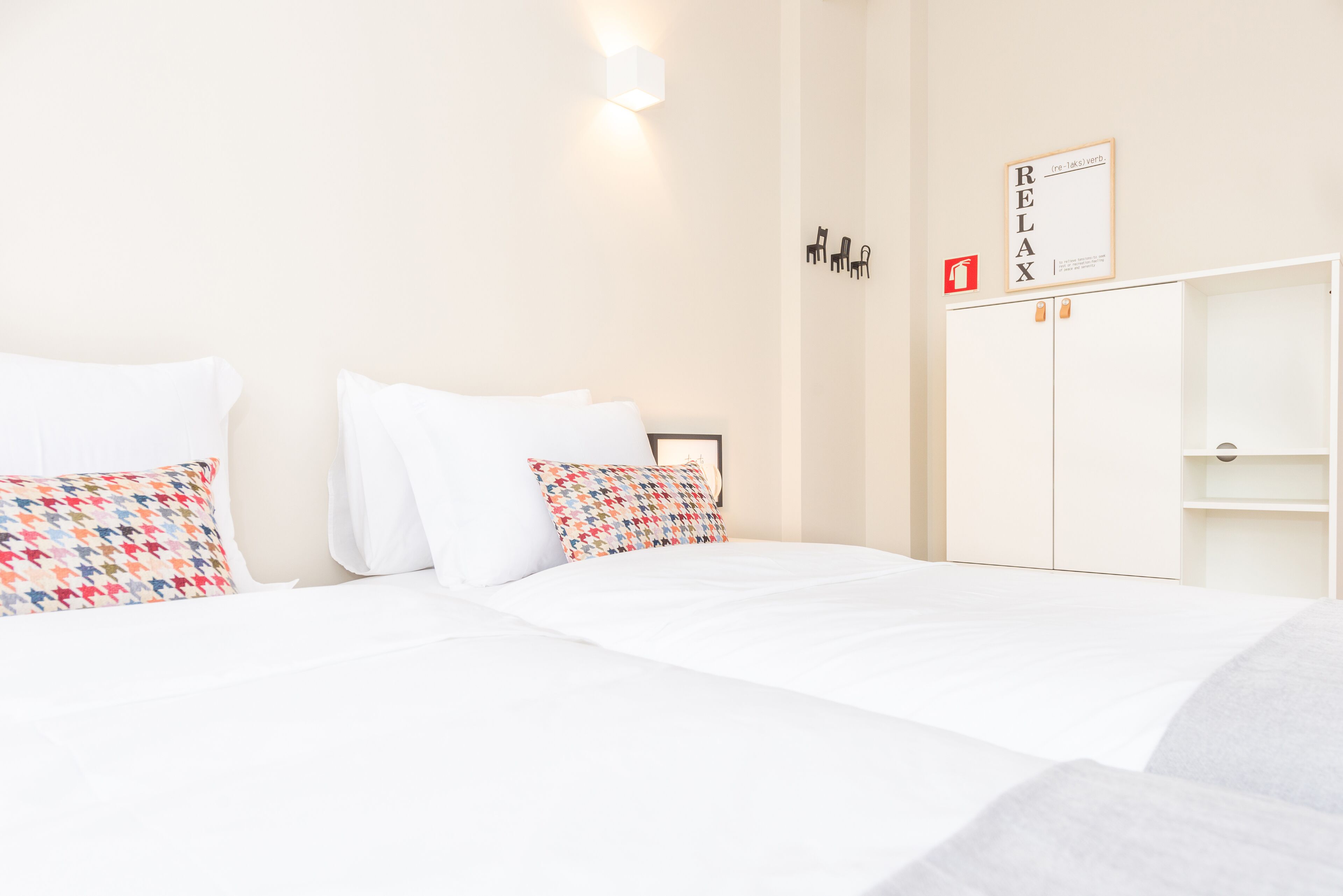 Et hotelværelse med en seng, hvidt sengetøj, en farverig pude med mønster, en vægmonteret lampe og et billede med ordet 'RELAX'.