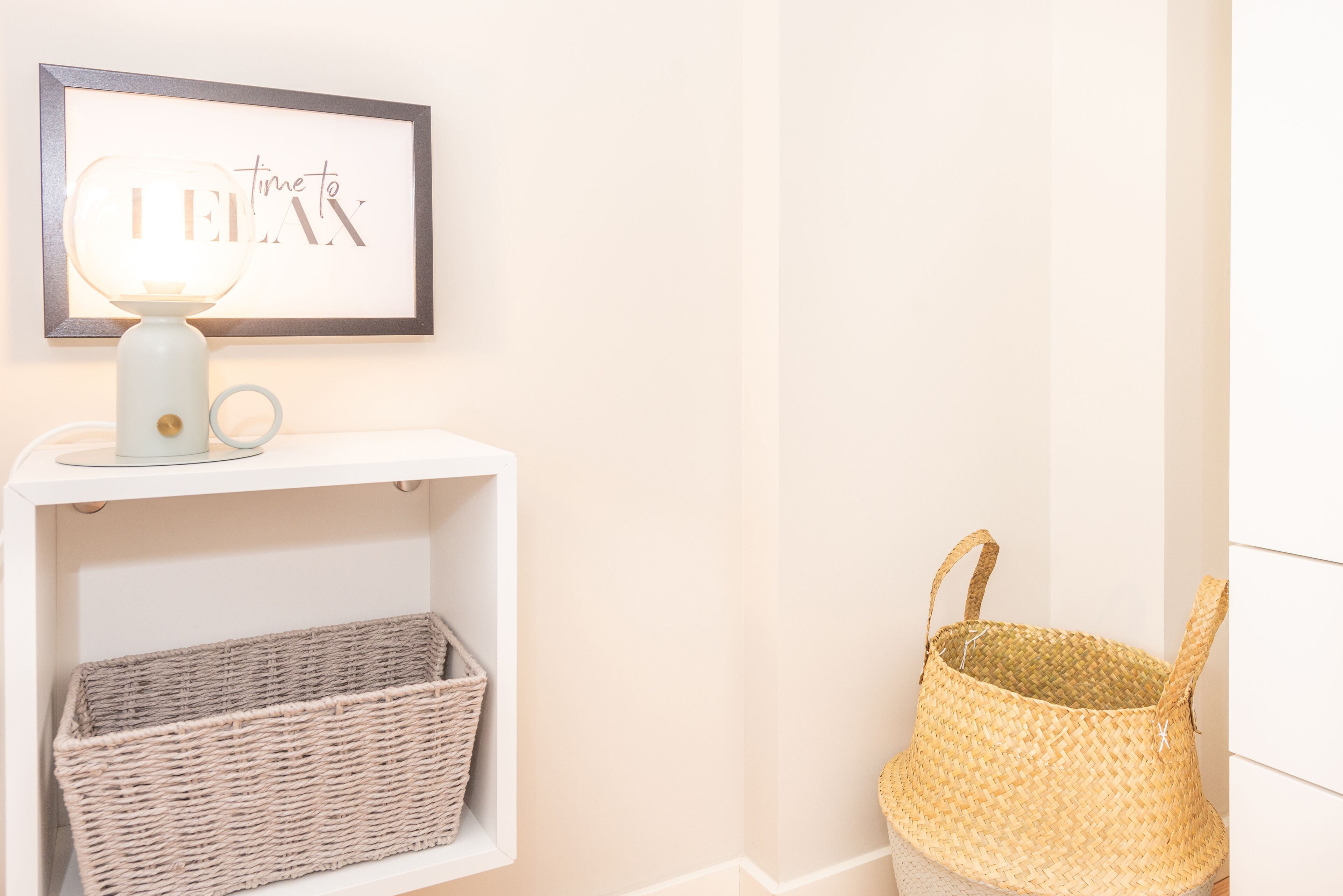 A framed artwork with the text "time to" on a wall, a white shelf with a wicker basket, and a woven basket on the floor.