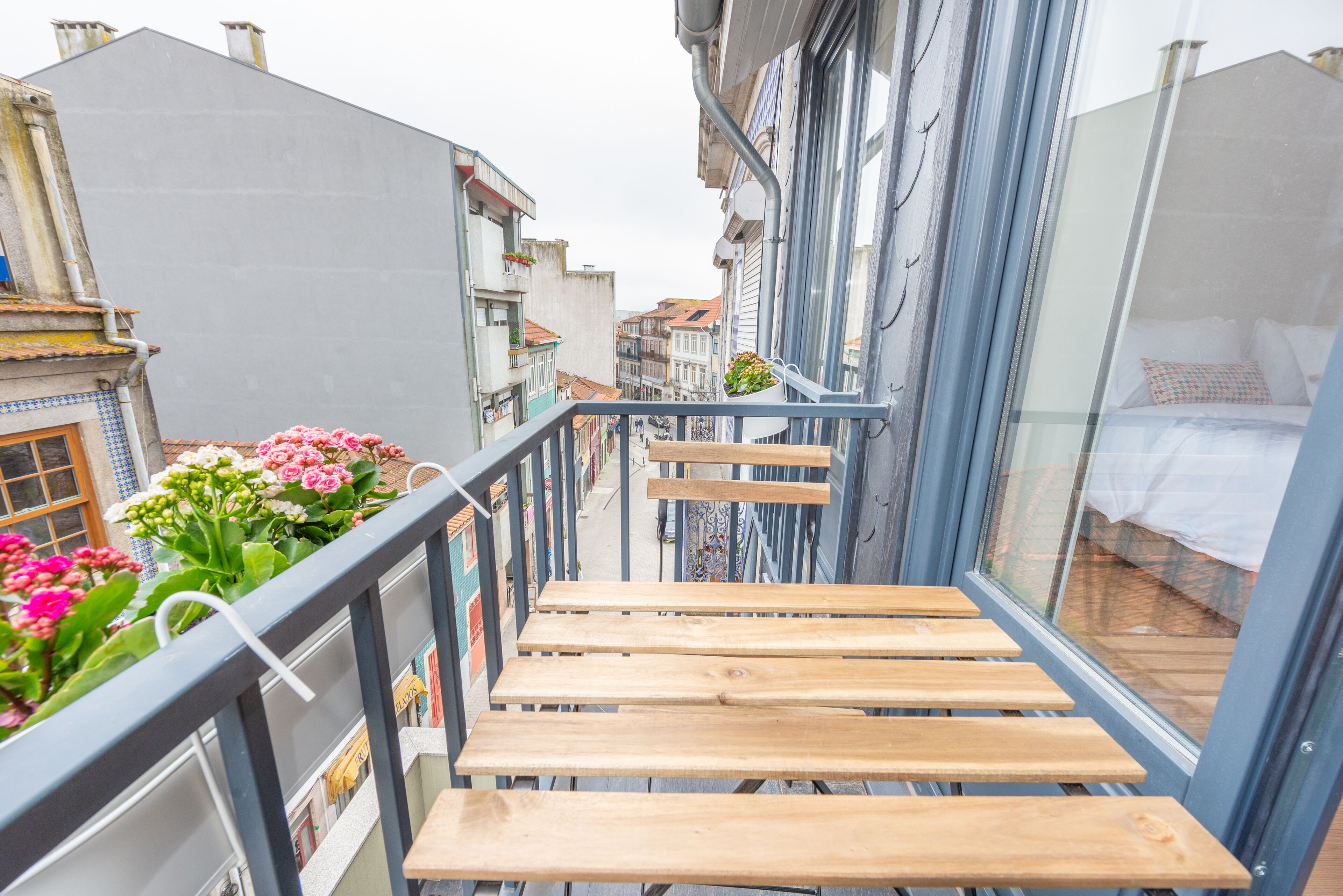 A narrow balcony with wooden steps leading to a bed, overlooking a street with buildings and flowers.