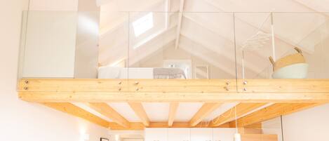 A modern loft bedroom with a glass ceiling, wooden beams, a kitchen area below, and a staircase.