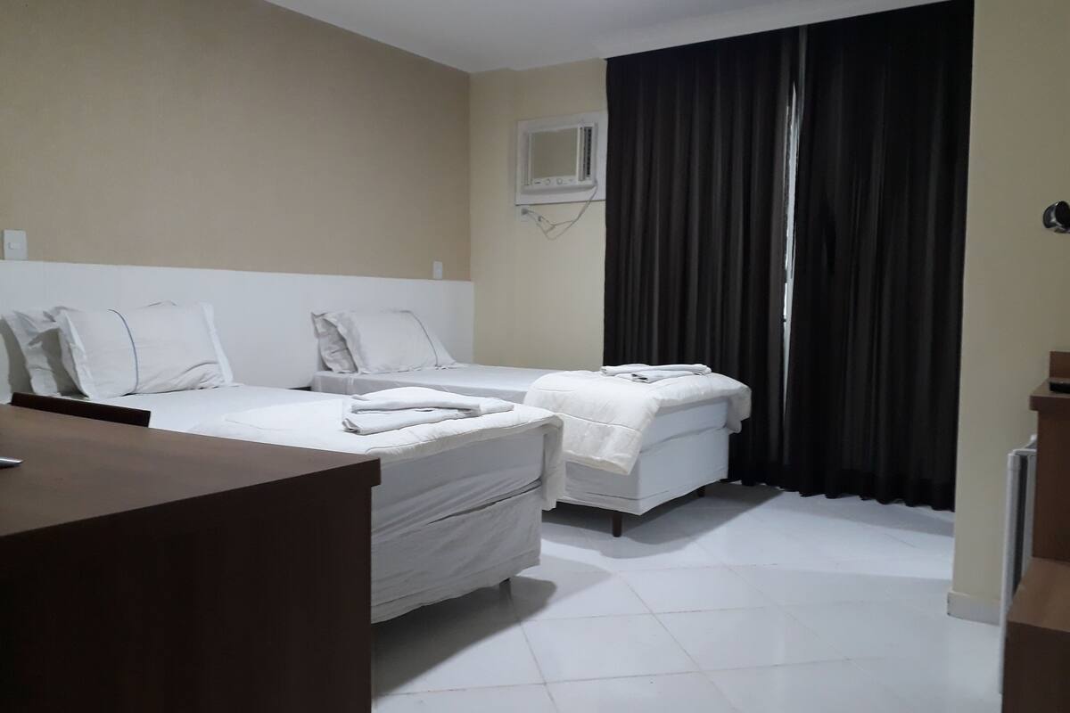A hotel room with two beds, a desk, a window with curtains, and a wall-mounted TV.