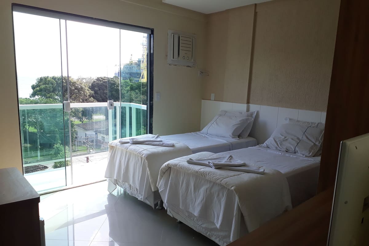 A hotel room with two beds, a large window with a view of greenery and construction outside, and a ceiling-mounted air conditioning unit.