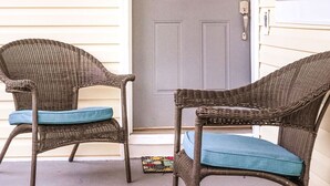 Une véranda avec deux fauteuils en osier dotés de coussins bleus, une porte grise ornée d’une guirlande et une fenêtre sur la droite.