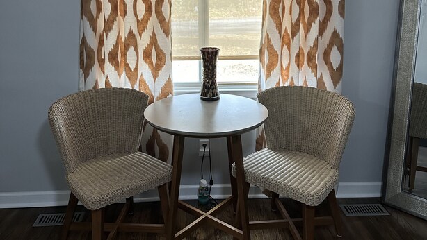 Une pièce avec deux fauteuils en osier disposés autour d’une petite table ronde, un vase sur la table et une fenêtre ornée de rideaux à motifs.