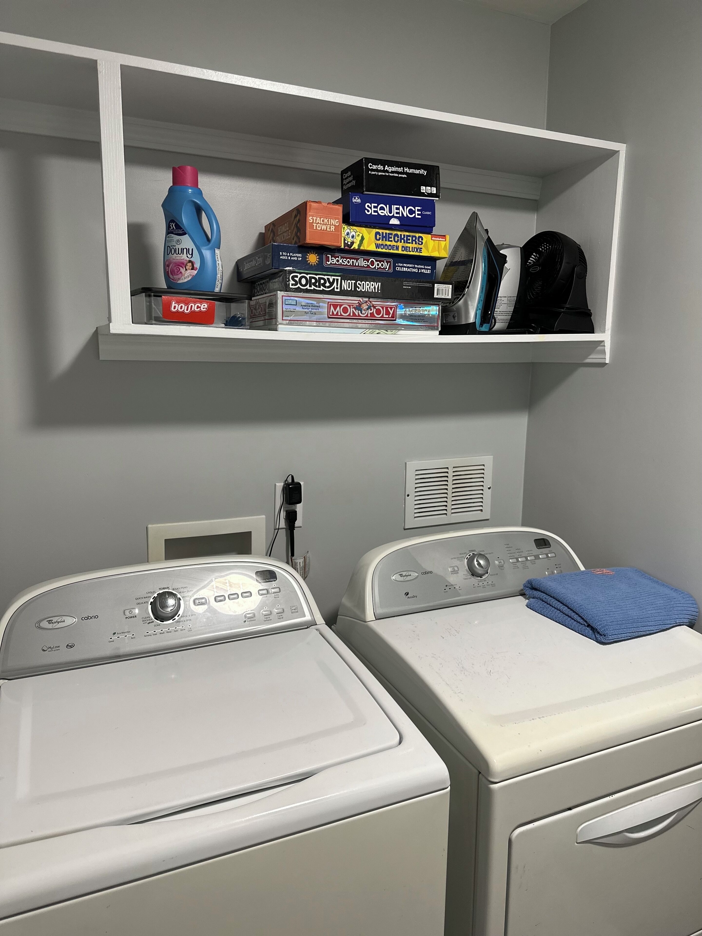 A laundry area with two front-loading washers and a shelf above containing various board games and a bottle of laundry detergent.