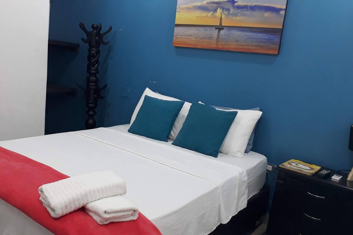 A neatly made bed with white and teal pillows, a red blanket, and white towels on a white mattress.