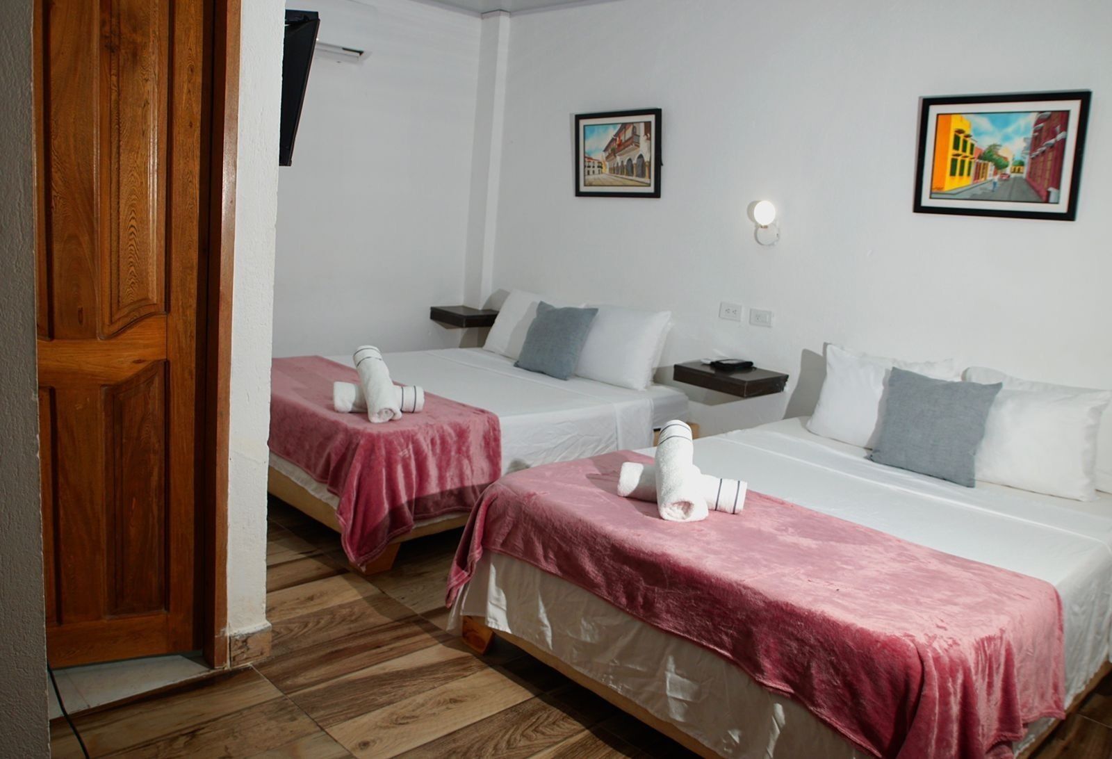 Una camera d'albergo con due letti, ciascuno con copripiumino rosa e cuscini bianchi. Si notano due quadri alle pareti, un comodino e una porta in legno.