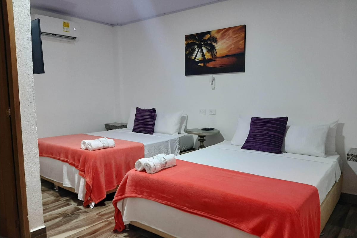 A hotel room with two beds, each with a red blanket and white pillows, a small table, and a painting of a sunset on the wall.
