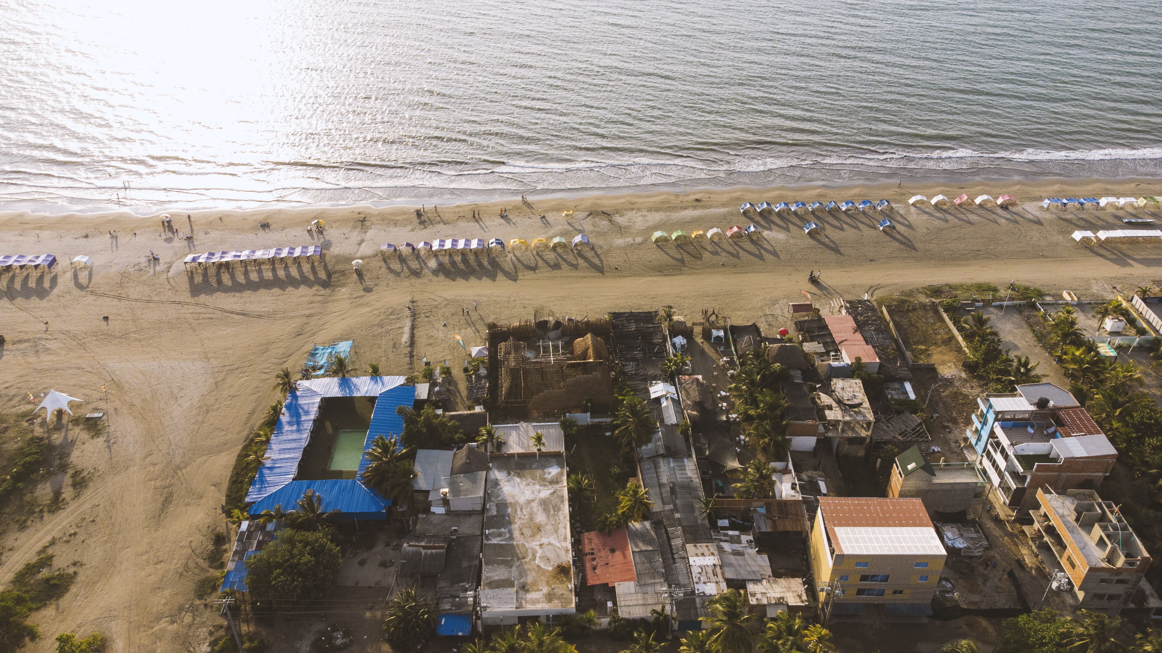 An aerial view of a coastal area with a beach, beach umbrellas, and a cluster of buildings with blue roofs.